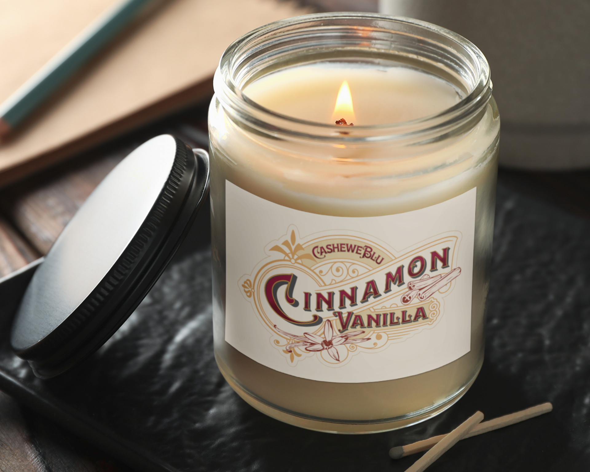 Cinnamon Vanilla lit 9 oz soy candle on table with used matches, notebook, coffee cup and lid leaning on jar