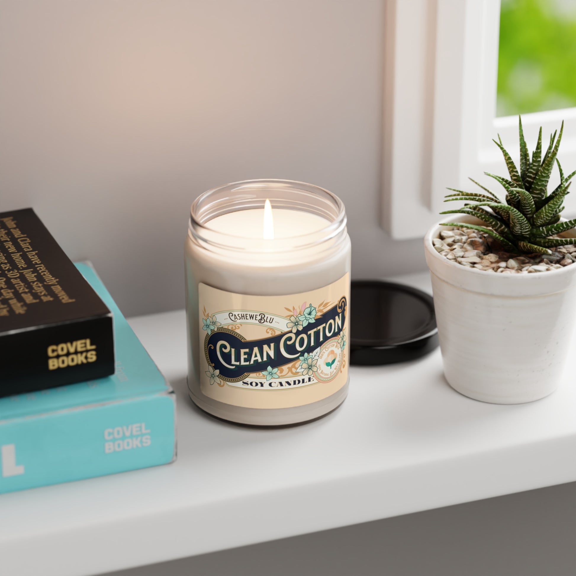 Clean Cotton 9 oz soy candle lit sitting on shelf next to books, plant and window