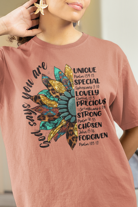 Terracotta color t-shirt with half a sunflower with gold, teal and leopard print petals and a teal middle with the words "God says you are" arching it on the left of the flower and the words listed with referenced bible verses "unique, special, lovely, precious, strong, chosen and forgiven" filling the the opposite side of the flower, worn by woman.