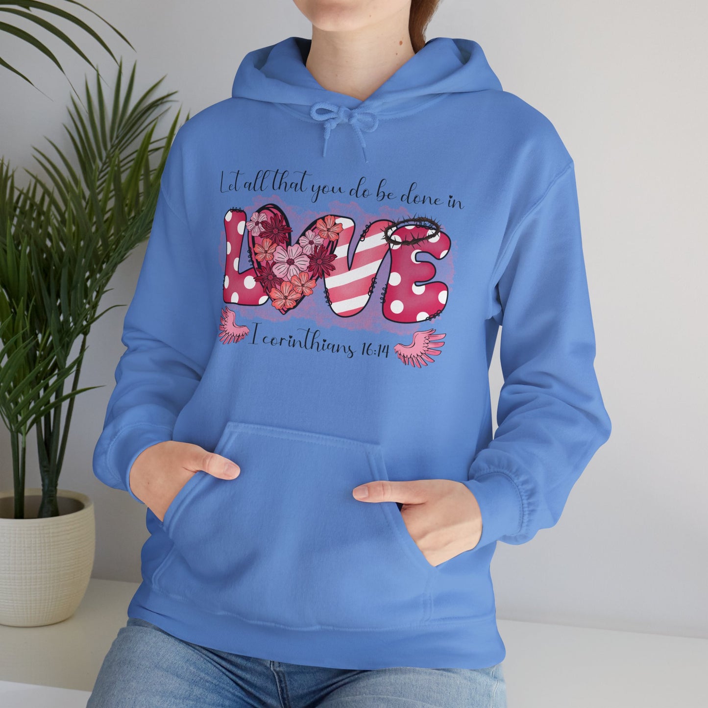 Carolina Blue hoodie with a playful and fun front "Love" graphic.  The word love is represent by white polka dots with pick back ground on the L and E and the E has a thorn crown.  The V is pink and white stripes and the O is represented by a heart filled with flowers.  Above in scripts says "Let all that you do be done in" and below the word love are wings flanking either side of I Corinthians 16:14. Modeled by Woman