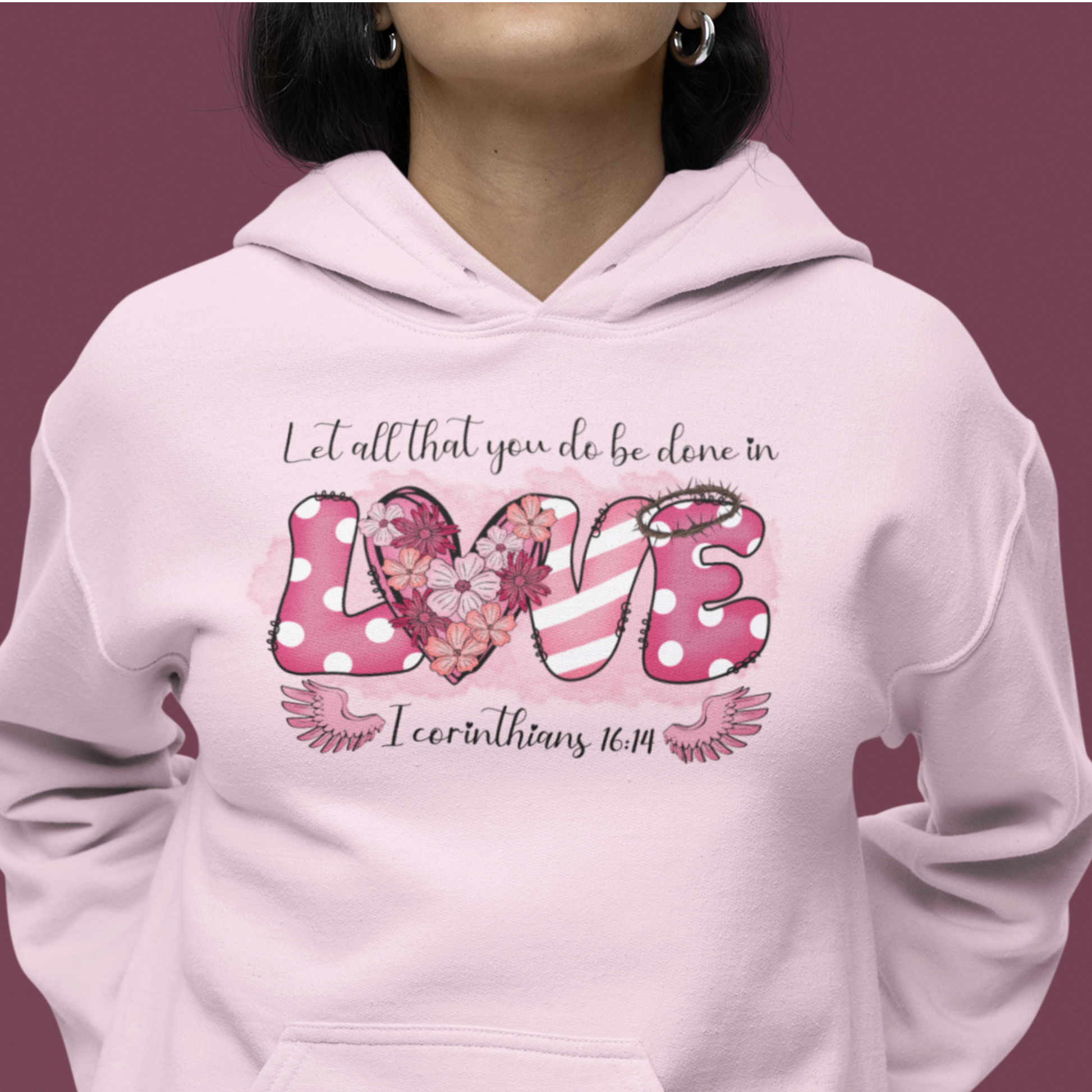 Light pink hoodie with a playful and fun front "Love" graphic.  The word love is represent by white polka dots with pick back ground on the L and E and the E has a thorn crown.  The V is pink and white stripes and the O is represented by a heart filled with flowers.  Above in scripts says "Let all that you do be done in" and below the word love are wings flanking either side of I Corinthians 16:14.  Modeled by woman.