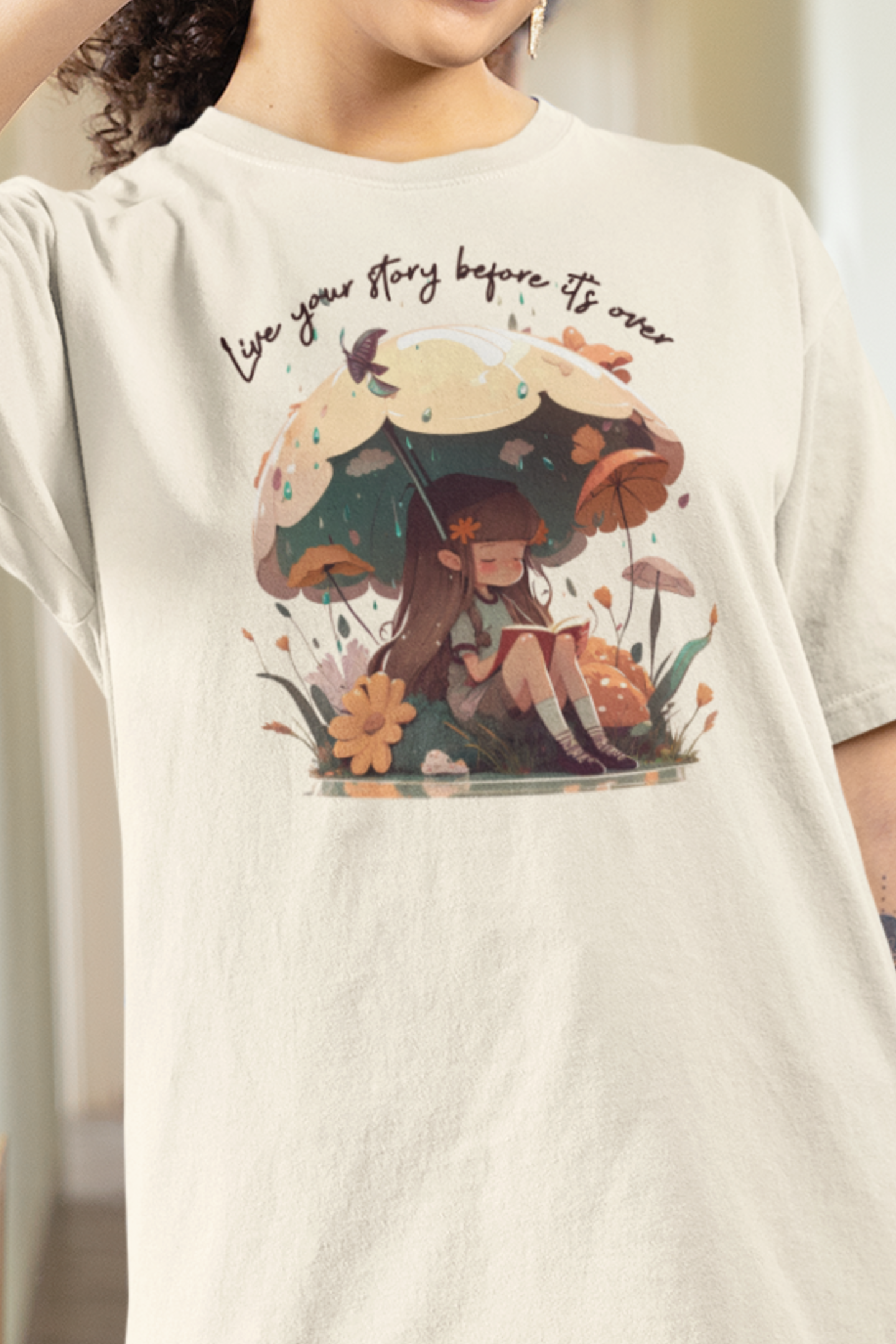 Ivory colored T-Shirt with girl sitting under an umbrella reading a book in the rain surrounded by flowers and mushrooms with the words live your story before it's over arching above the umbrella