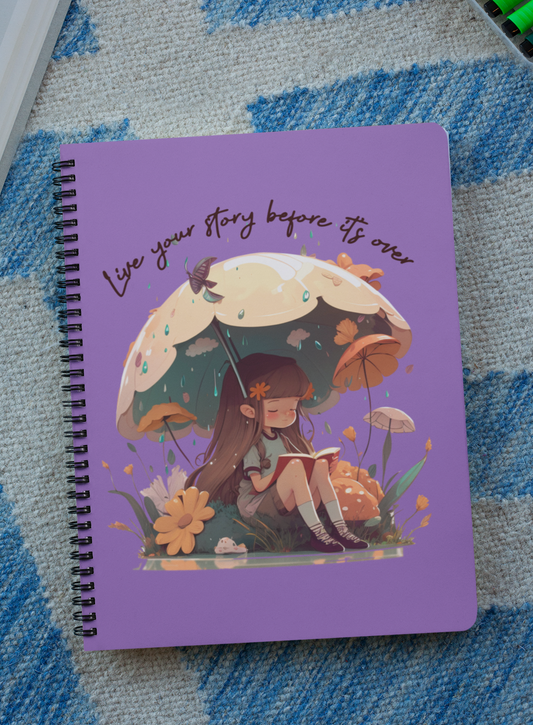 Spiral notebook with girl sitting under an umbrella reading a book in the rain surrounded by flowers and mushrooms with the words live your story before it's over arching above the umbrella