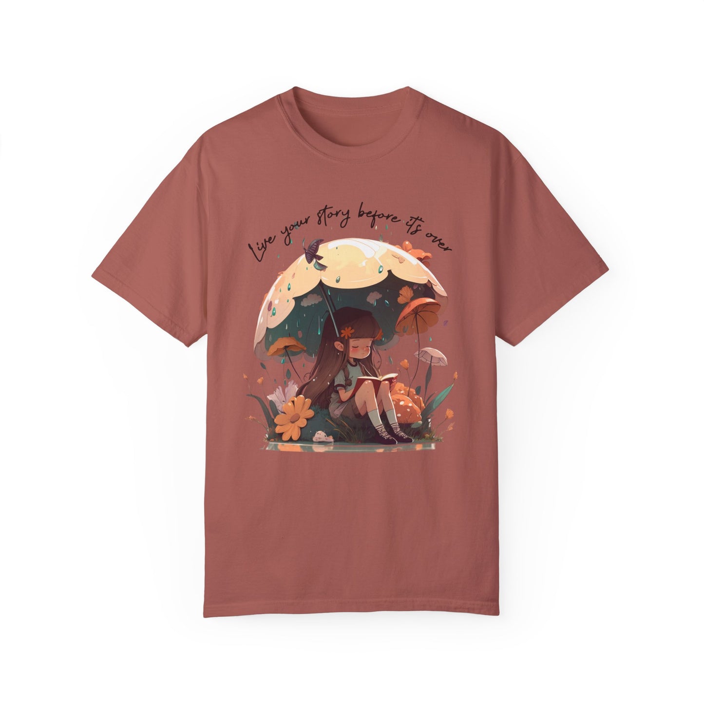 Cumin colored T-Shirt with girl sitting under an umbrella reading a book in the rain surrounded by flowers and mushrooms with the words live your story before it's over arching above the umbrella