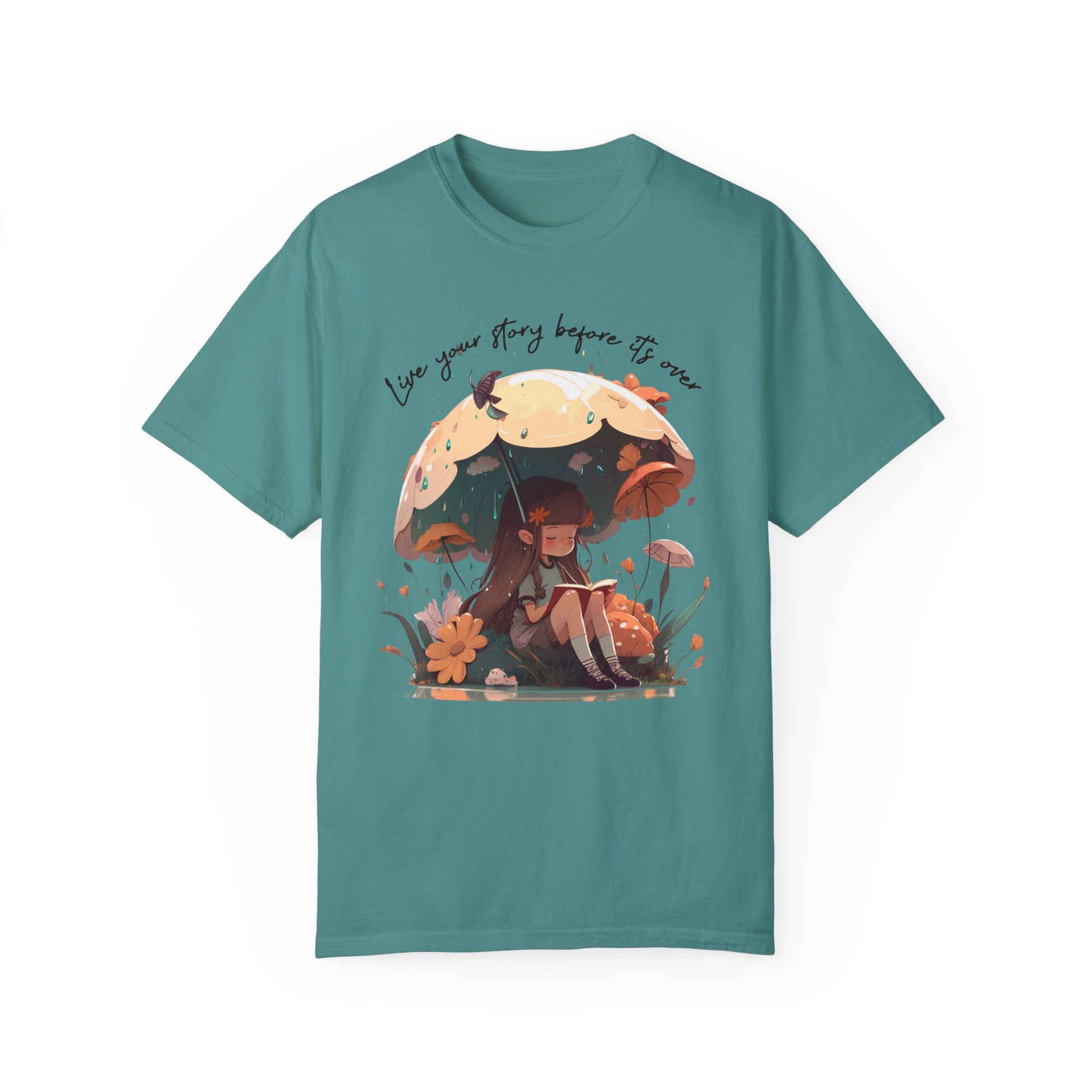 Seafoam colored T-Shirt with girl sitting under an umbrella reading a book in the rain surrounded by flowers and mushrooms with the words live your story before it's over arching above the umbrella