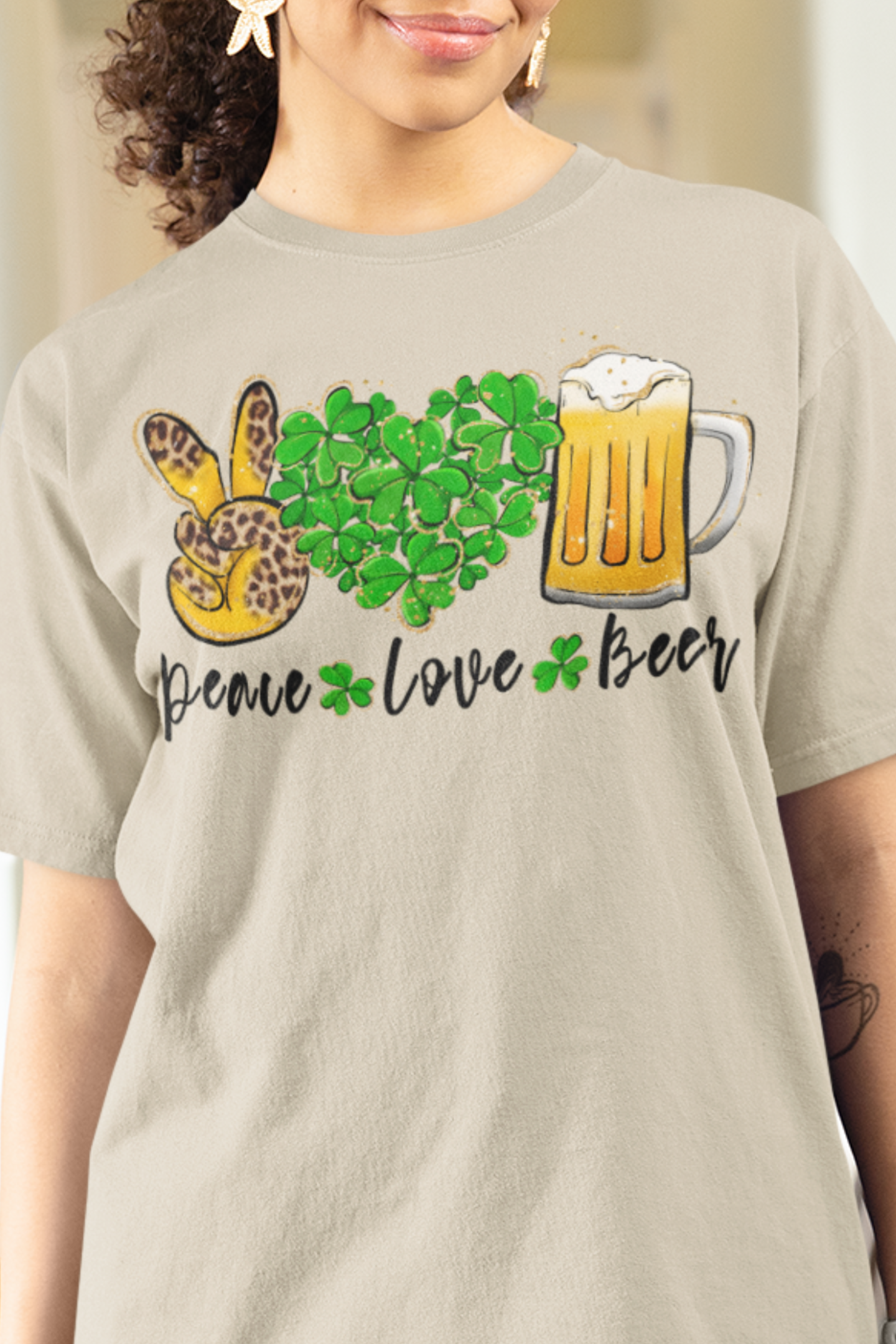 Sand Color T-Shirt with the words Peace, Love, Beer written on it with a hand in a peace sign and heart filled with shamrocks and a mug of beer above each of the words. Mock-up woman